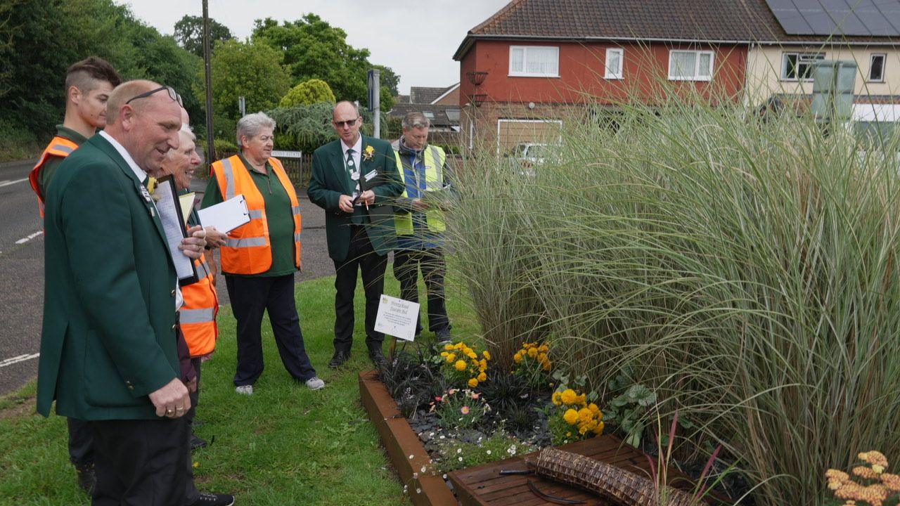 Judges in dark green waistcoats carrying clipboards assessing a floral display in Chatteris. The main feature is a clump of tall grass with smaller flowers dotted around it.