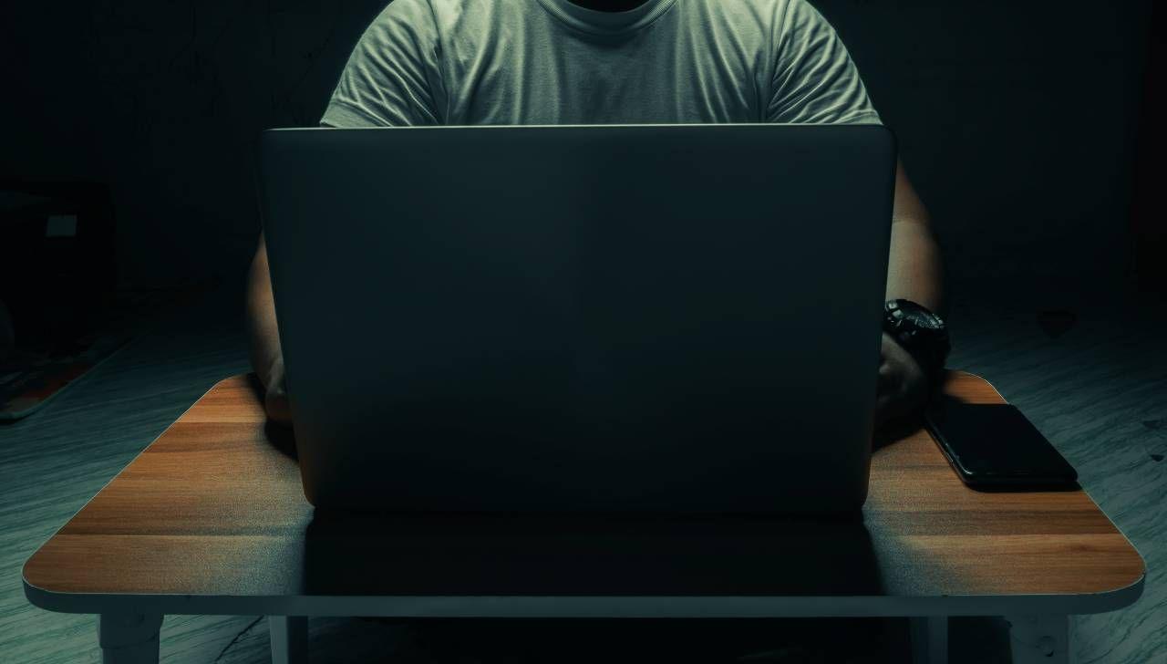 An anonymous man sits behind a laptop. The image is cropped so you cannot see the man's head. The laptop sits on a wooden table with a phone also sat next to the computer.