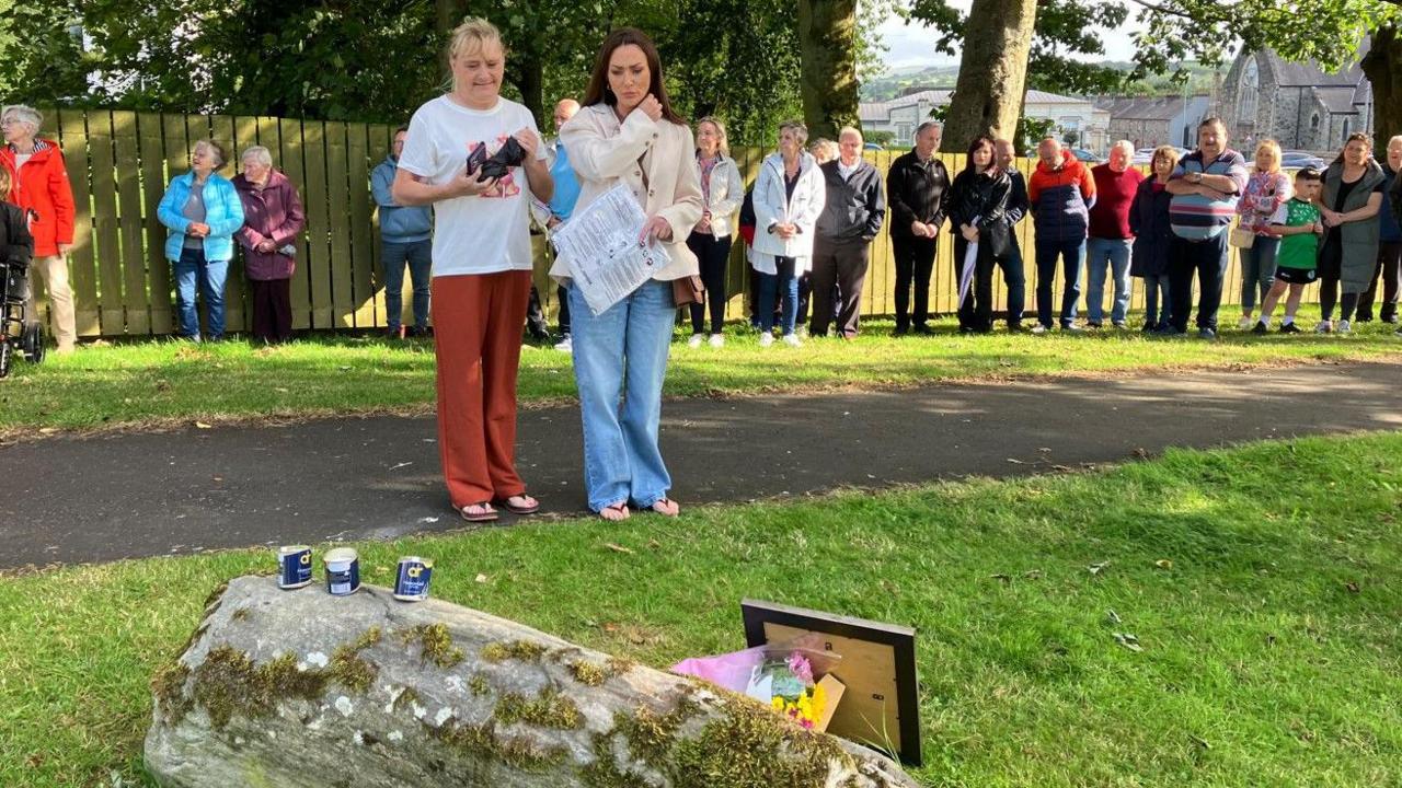 Two people stand and look at the memorial stone, with others looking on behind them.