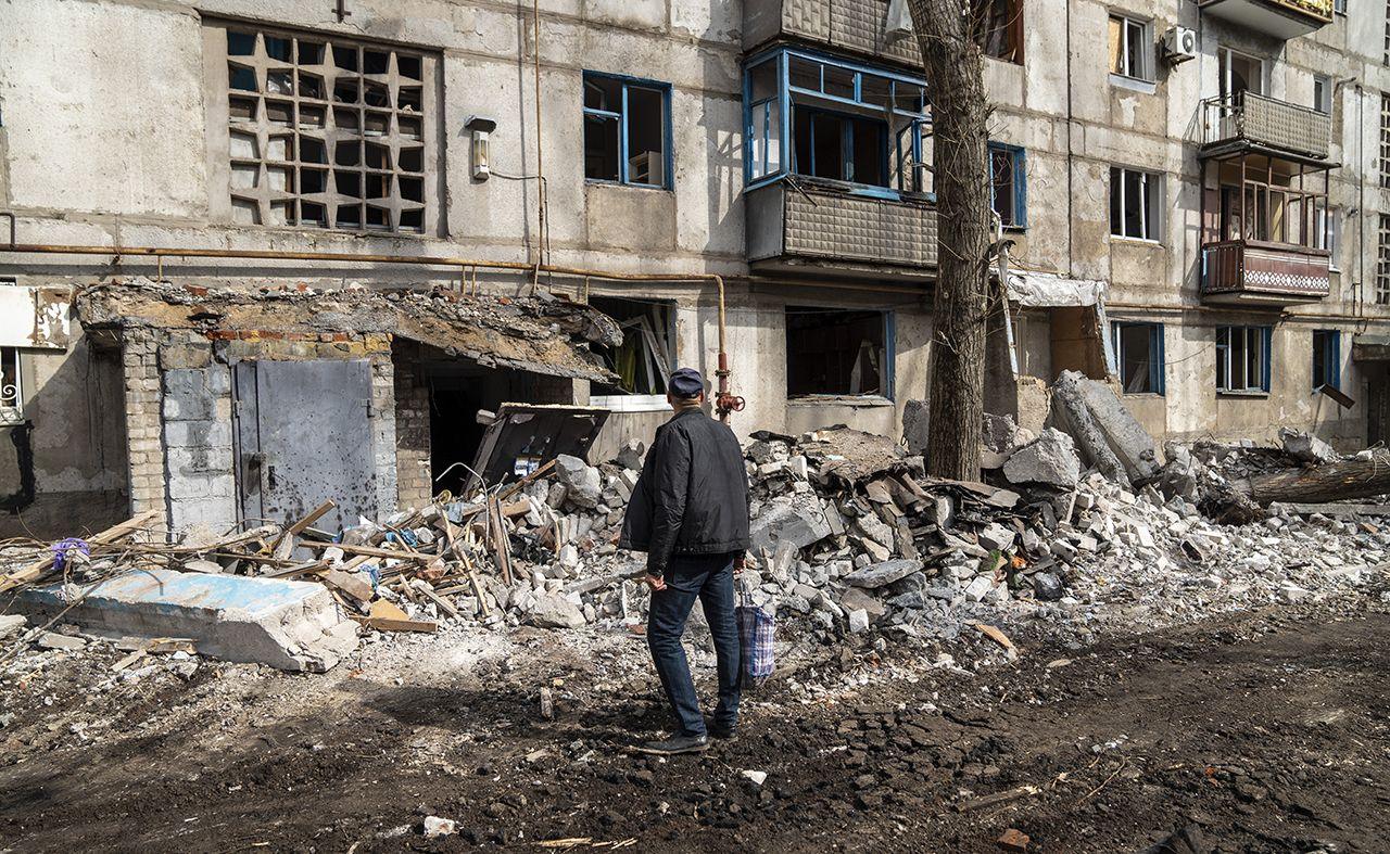 A person looks towards the rubble of an apartment block on a street in Kostiantynivka after Russian shelling on 13 March. They're wearing a cap and looking away from camera, and mud is seen on the street next to the pile of rubble with the building in the background.