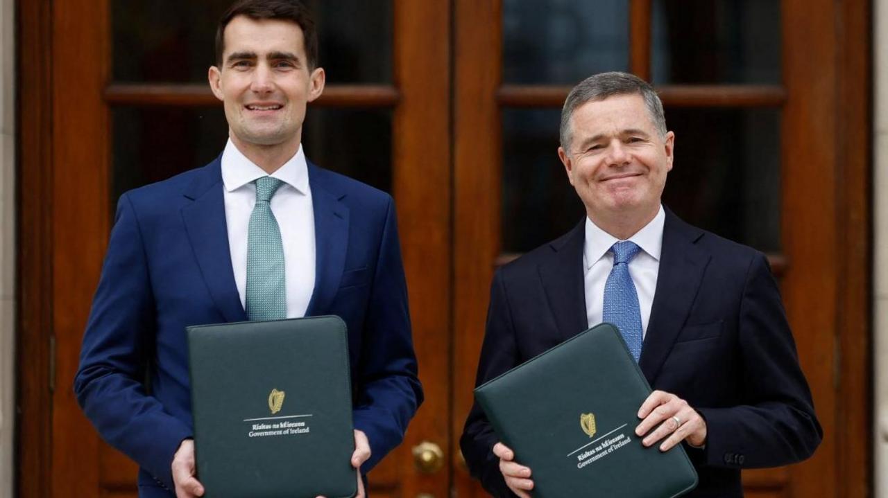 Jack Chambers, with dark hair, in blue suit, with white shirt and green tie, with budget document, along with Paschal Donohoe, who has grey hair. in navy jacket with white shirt and blue tie.