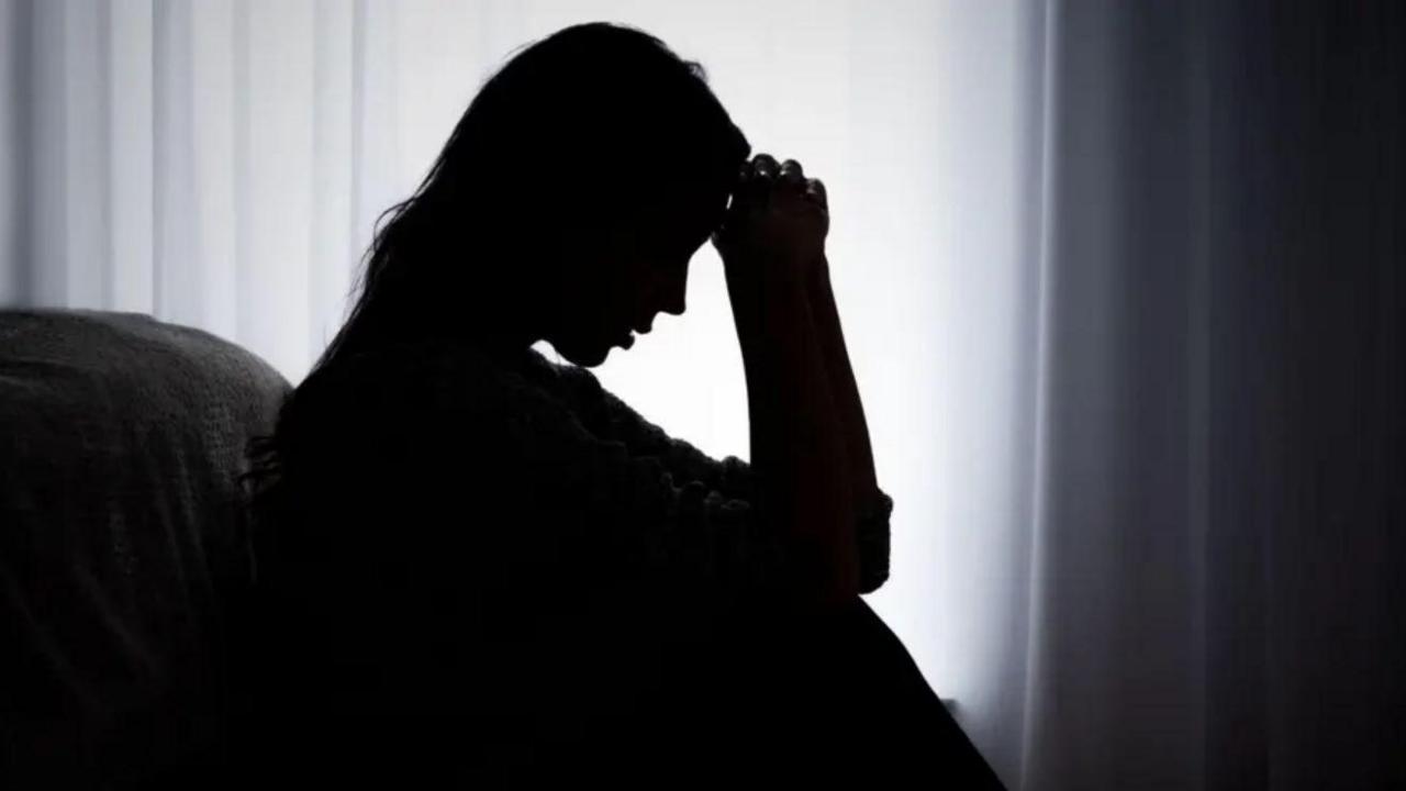 Shadow of a woman sitting down with her hand on to her head leaning against the foot of a bed. A white voile curtain is behind her with light coming in from a window. 