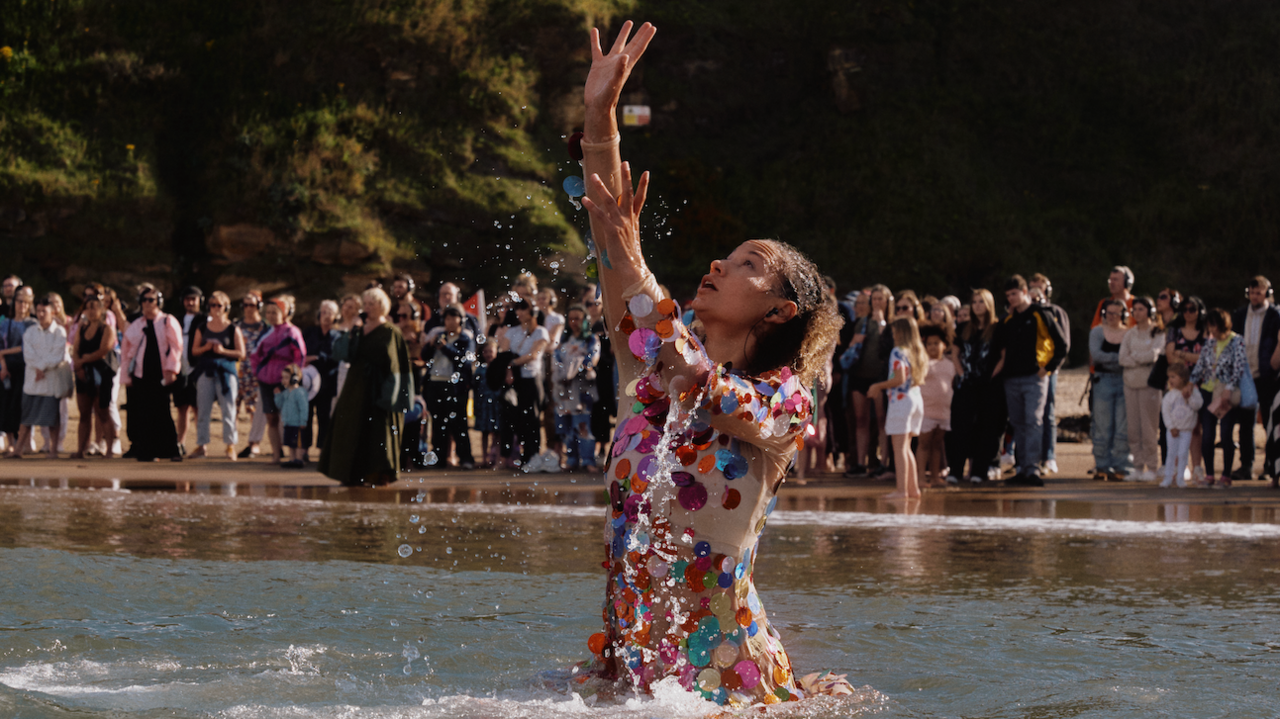 A woman performs a dance in the sea in front of a crowd of people gathered on the beach.