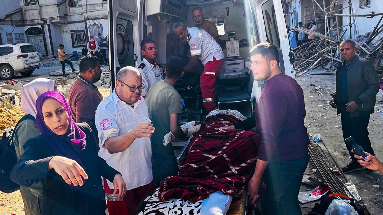 A group of people and medics, including people wearing shirts with the Palestinian Red Crescent logo, lift a wounded patient on a stretcher into the back of an ambulance on a street, with damage and debris in the background, outside the Kamal Adwan Hospital in northern Gaza on 28 October