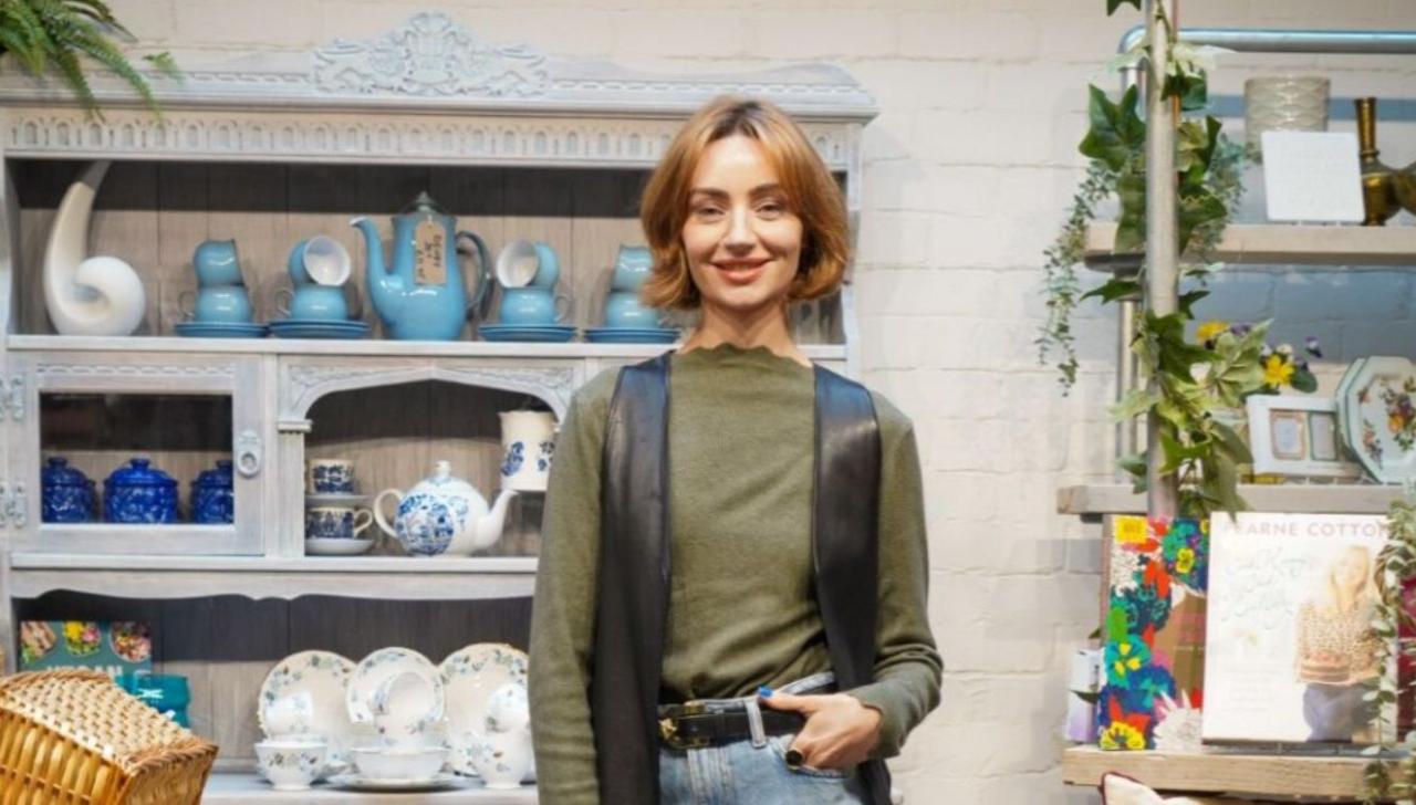 Jen Graham, who is wearing a long-sleeved cotton jumper and black leather waistcoat, blue jeans and a black belt, is smiling at the camera on this posed shot. She is standing in front of a cabinet of crockery.