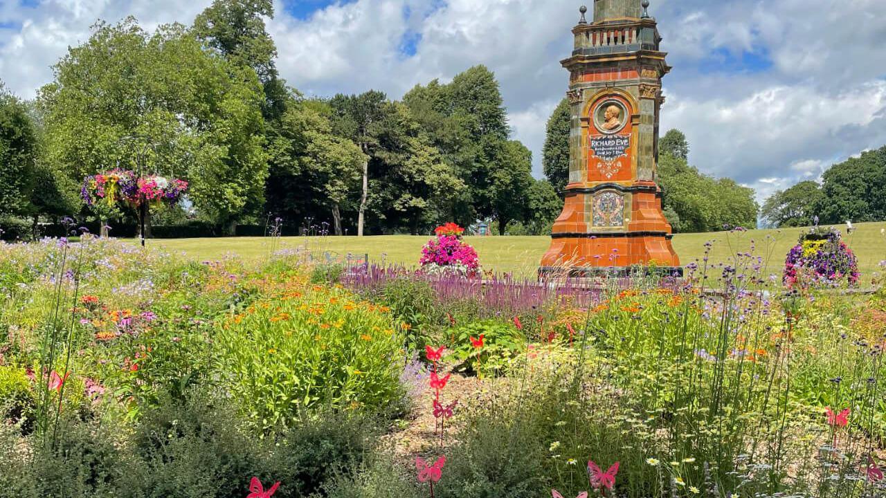 A garden with pink, red and purple flowers throughout. There are small red metal butterflies around the garden while trees can be seen in the background 