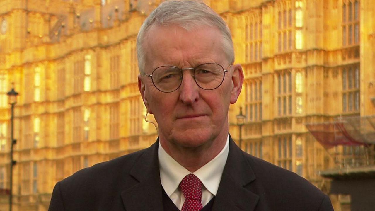 A man look at the camera, he has short grey hair and round glasses. He is wearing a suit with a red tie and white shirt and dark overcoat. He is standing outside in front of the Houses of Parliament in London. 