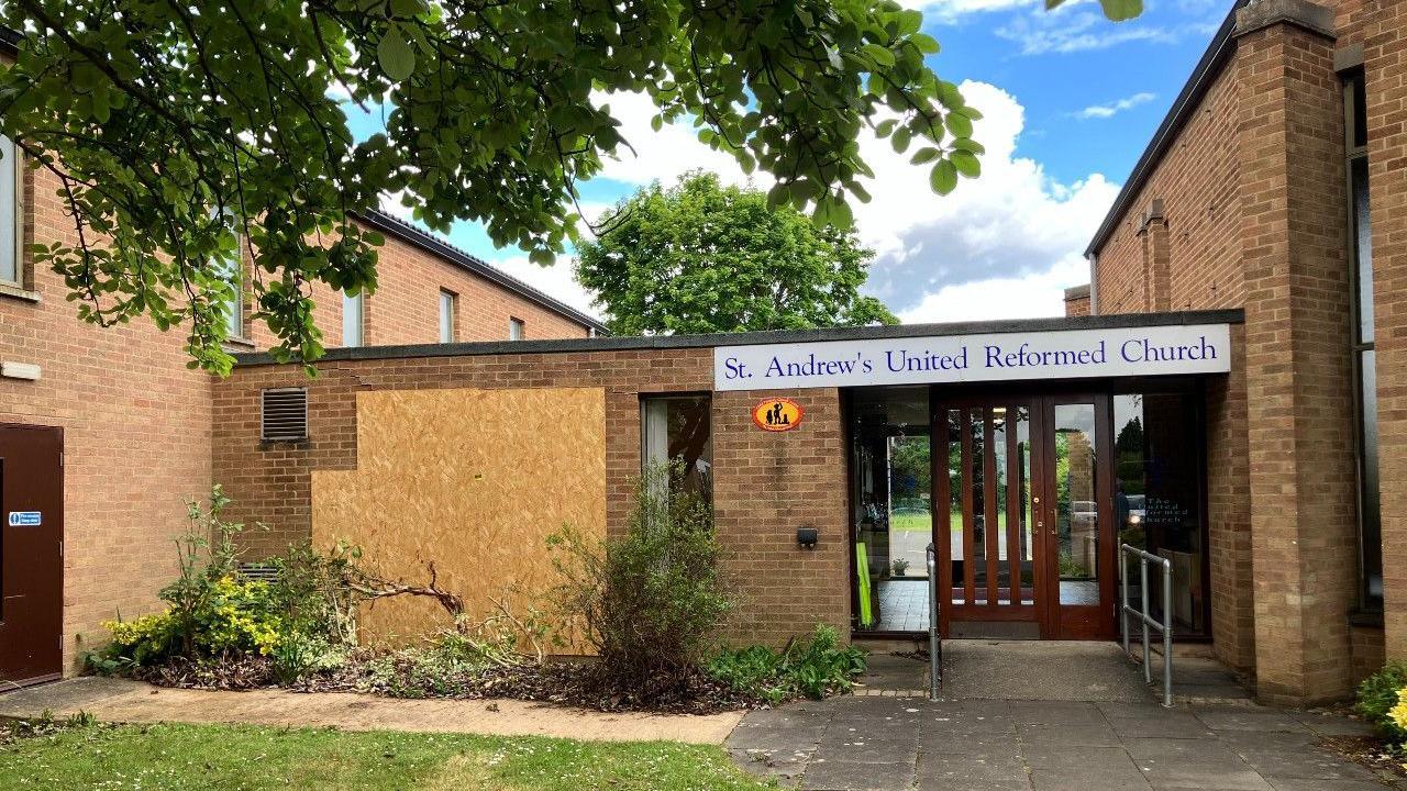 Exterior of the church building- a brick building with a brown glass door- and a boarded up wall on the left with damaged trees in front of it