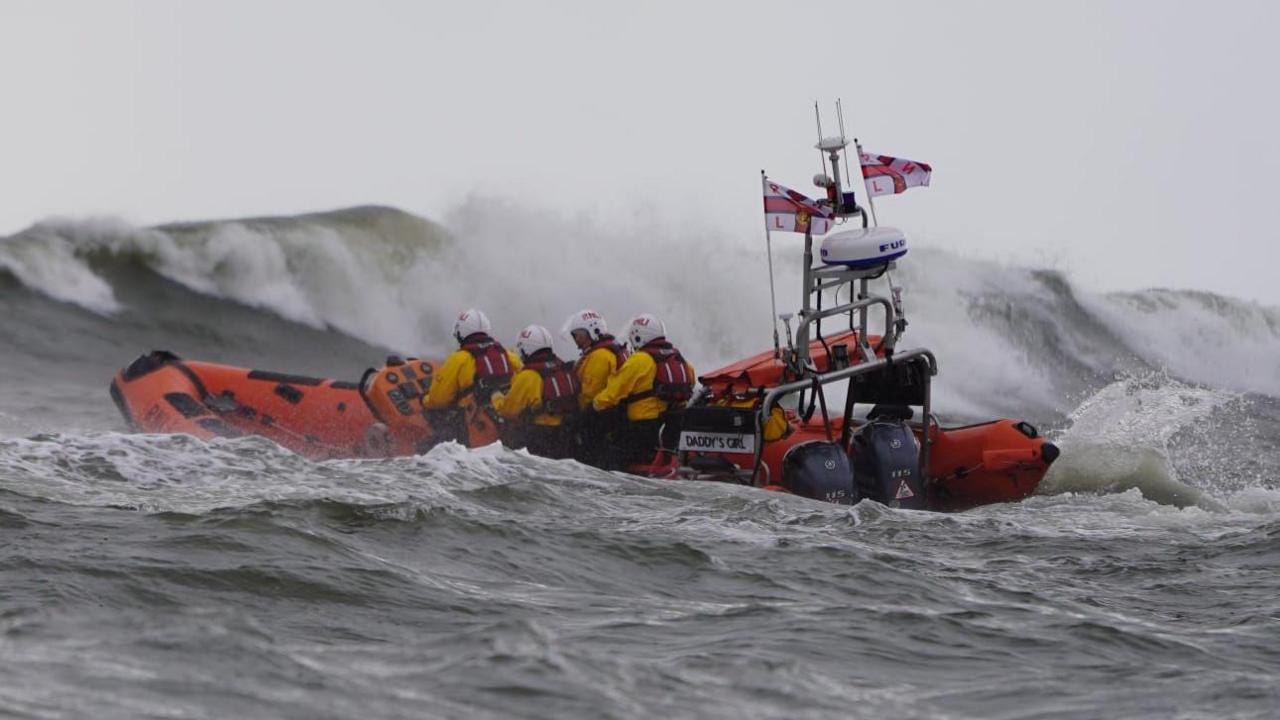 An orange lifeboat heads out to sea among large choppy waves. It is an open top boat with inflatable sides and there are four crew aboard each wearing a yellow drysuit, orange lifejacket and white helmet