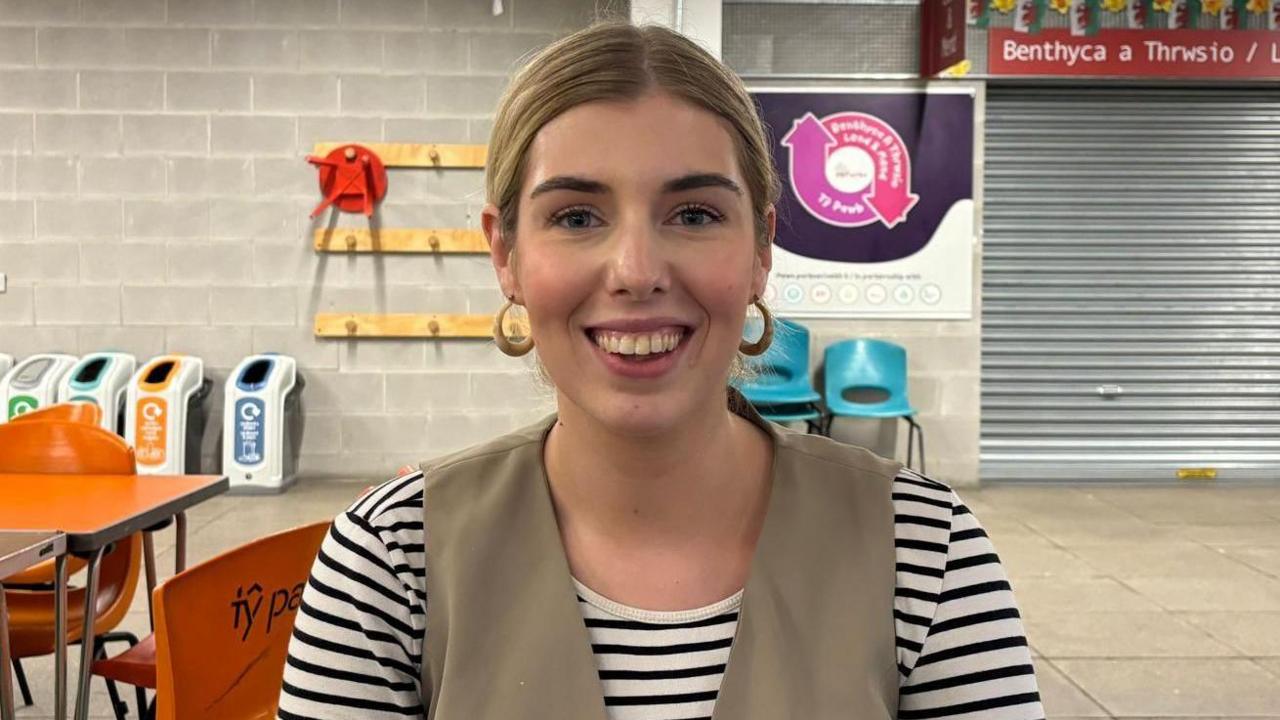 A blonde woman with hoop earrings and a stripy top sat down smiling at the camera. It is a head and shoulders shot of her. 