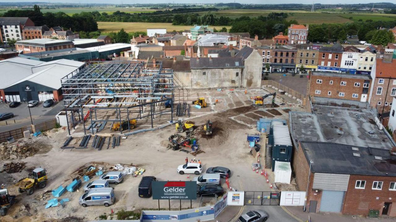 A construction site in a town centre. Steel framework for a large building is partially built, with machinery, vehicles, and workers scattered around. Surrounding the site are shops, older buildings, and open countryside in the distance.