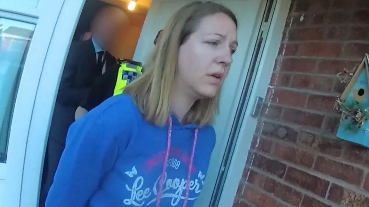 Police body-cam footage of Lucy Letby being led out of her front door by officers, wearing a blue hoodie with her hands behind her back. 