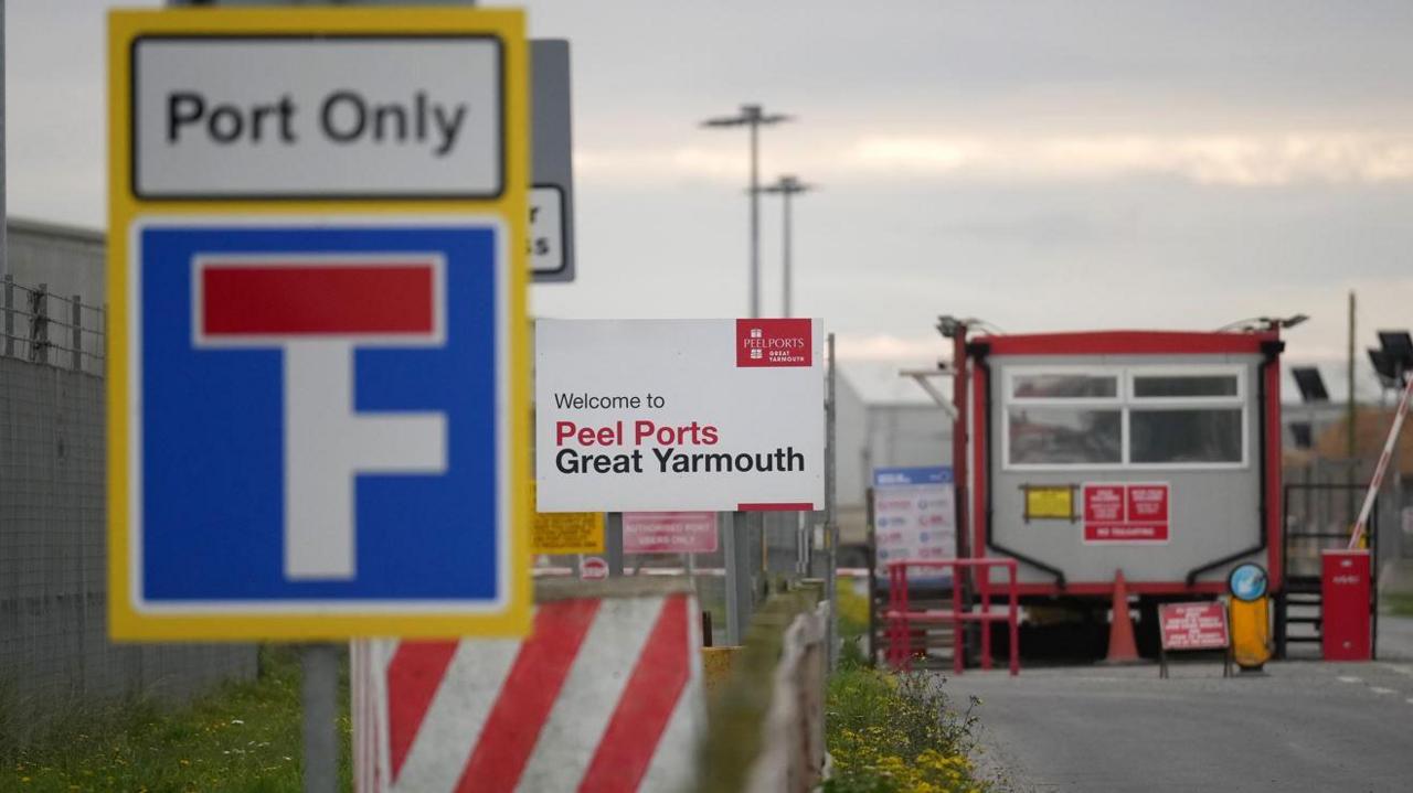 Front gate of a port facility showing signs, barriers and a security hut