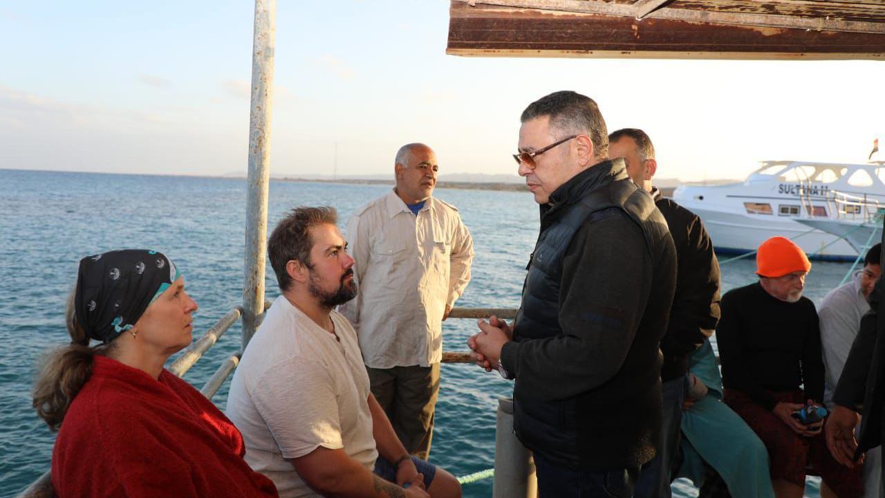 Red Sea Governor Maj-Gen Amr Hanafi (in the centre) standing with hs hands clasped, wearing a black gilet and sunglasses, as he talks to survivors of the boat sinking (on the left). There is a woman wearing red with a black bandana next to man in a white t-shirt. There are also other men behind Hanafi. In the background is the sea with a boat.