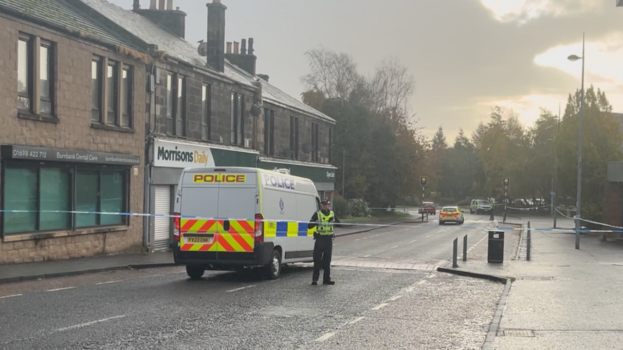 A street scene in low autumn light showing a police Transit van with officer alongside parked on the street. The street is cordoned off with tape and on the right a hooded man is using a cash machine