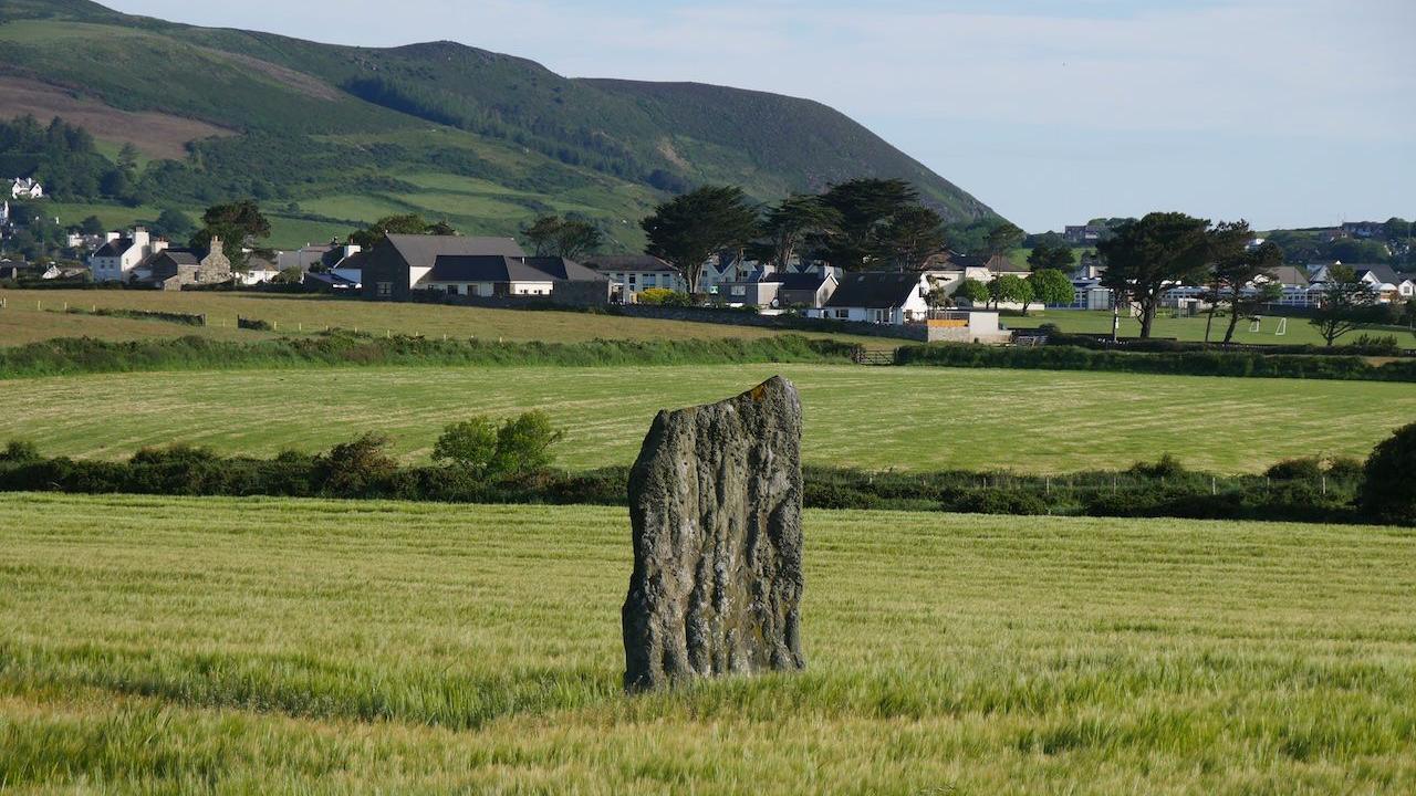 A large rock is in the middle of a field, you can see a village and hills in the background.