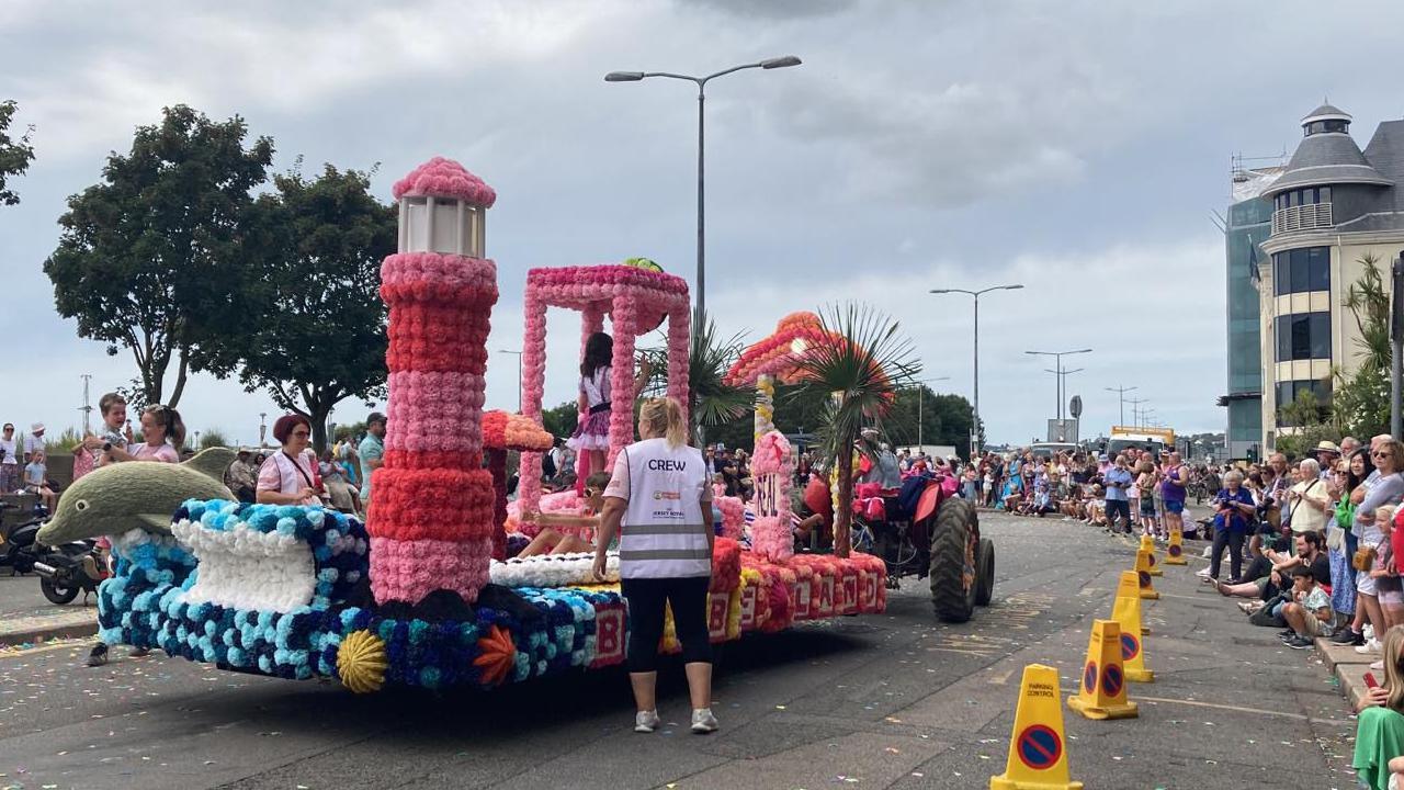 A largely pink coloured float which has a tall lighthouse on one corner and a dolphin leaping out of the other corner. There is a pergola  covered in pink flowers in which a young girl stands wearing a pink dress. At the front there is a real palm tree. Along the side of the float reads BARBIE LAND. There are many spectators lining the street.