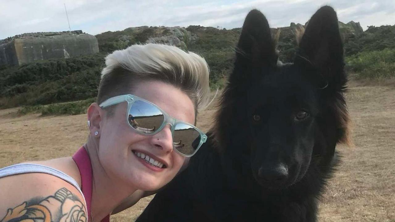 Stephanie Stefani on the left, wearing grey sunglasses, in a grey tank top, with short hair. She has a tattoo on her shoulder. Next to her is a black german shepherd. In the background is a field and grass. 