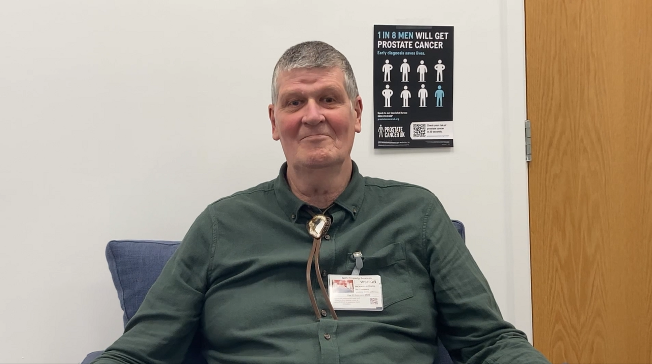 Richard wears a green shirt and is sitting in a hospital ward besides a poster highlighting the main symptoms of prostate cancer.