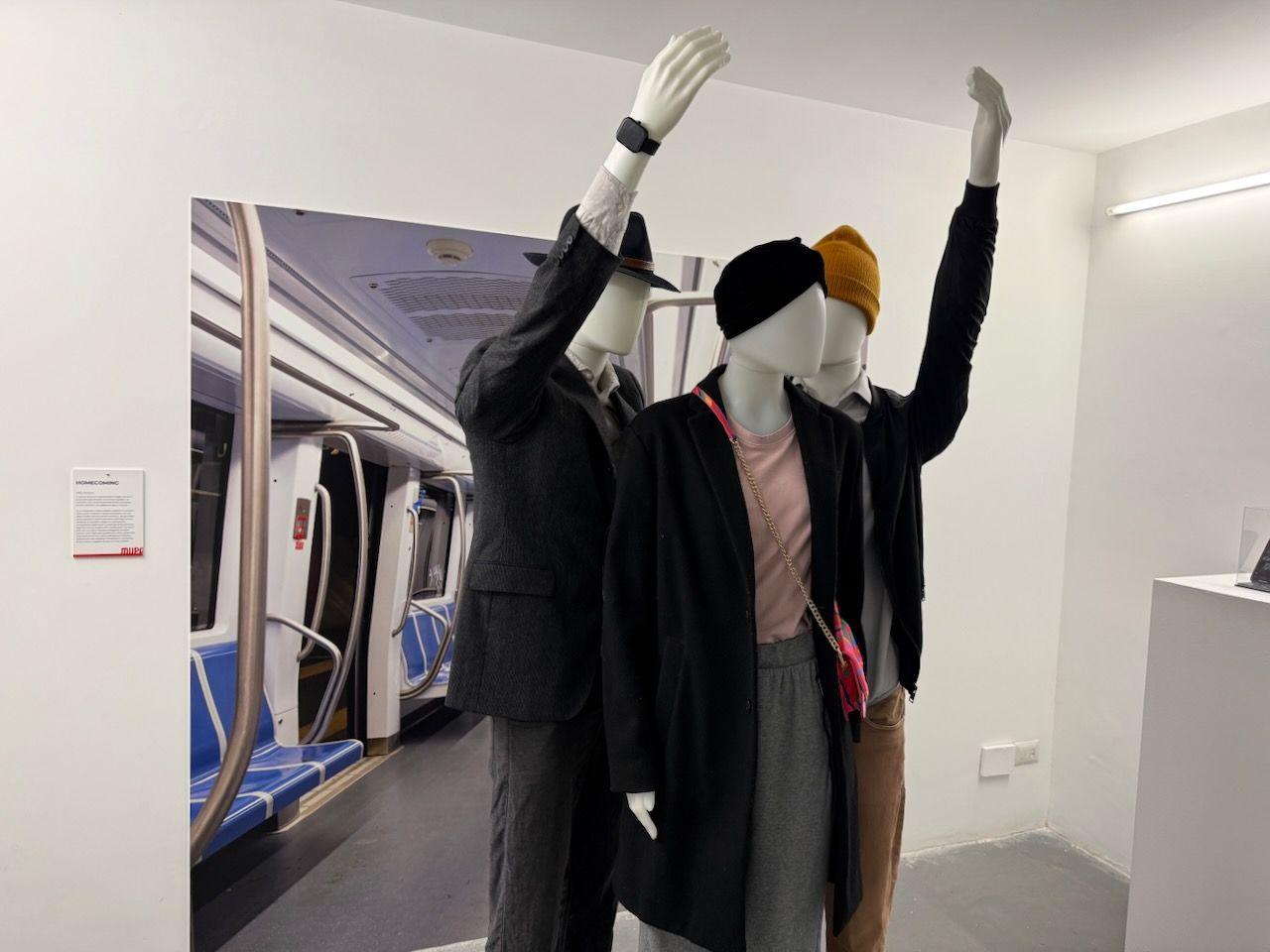 A plain white room with three dummies. One is dress as a woman and two as men. They are posed like they are posed as if they are riding the underground or metro with a display of a carriage behind them.
