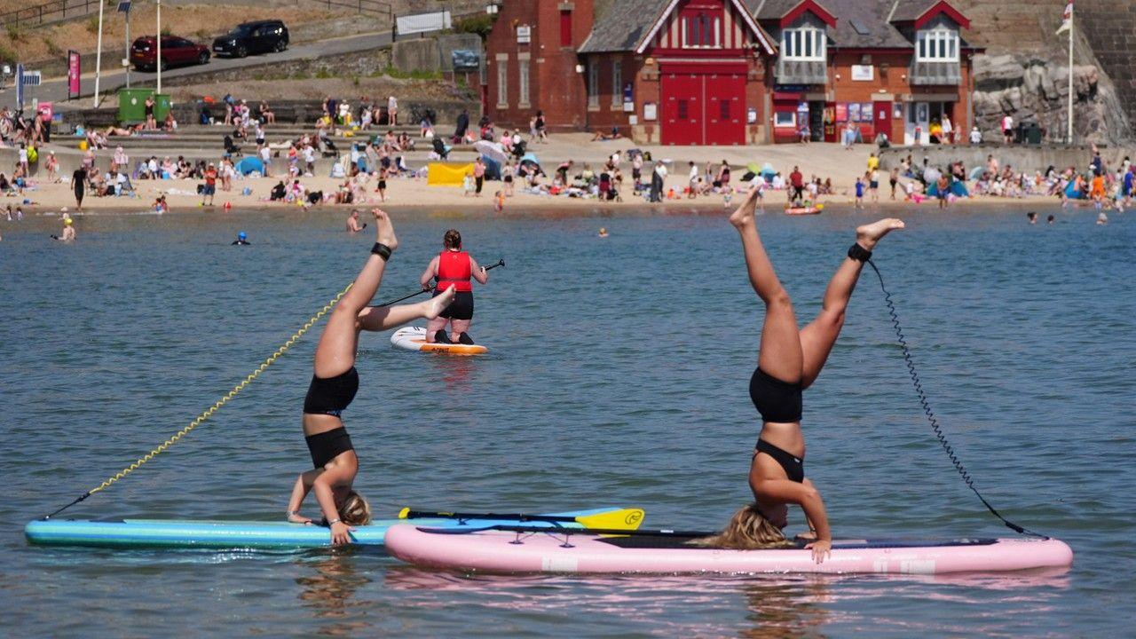 Two women stand on their heads on paddle boards in the sea, with people in the distance on the beach
