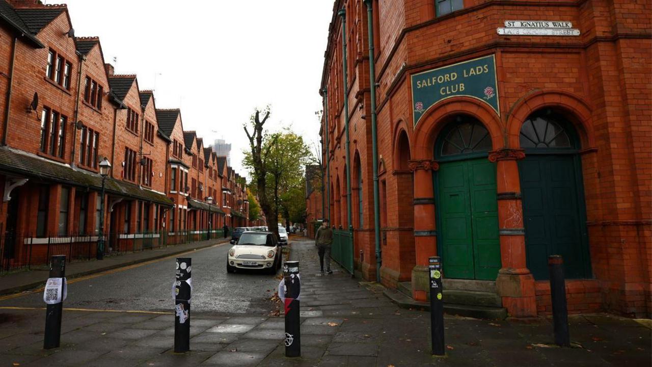 A street view of the outside of Salford Lads Club, a red-tiled building with a green sign with embossed gold lettering. A man can be seen walking passed on an overcast day.