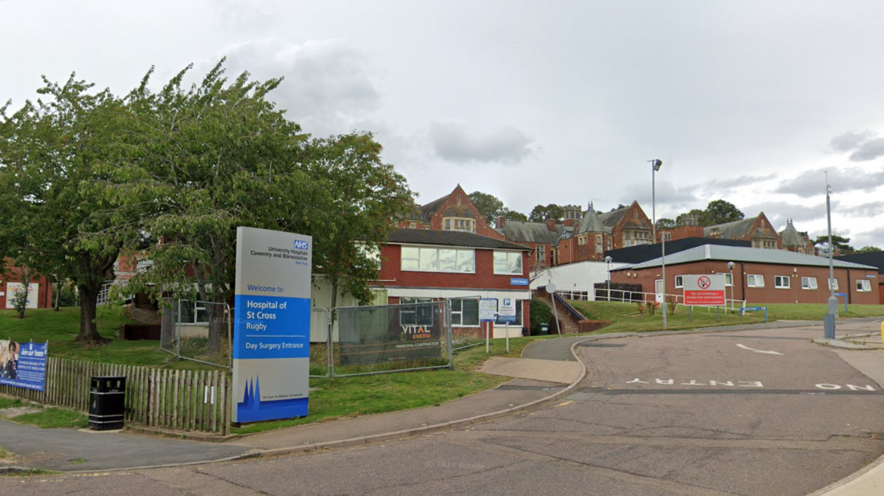 The front of the Hospital of St Cross with a blue and silver NHS sign and brick buildings in the background