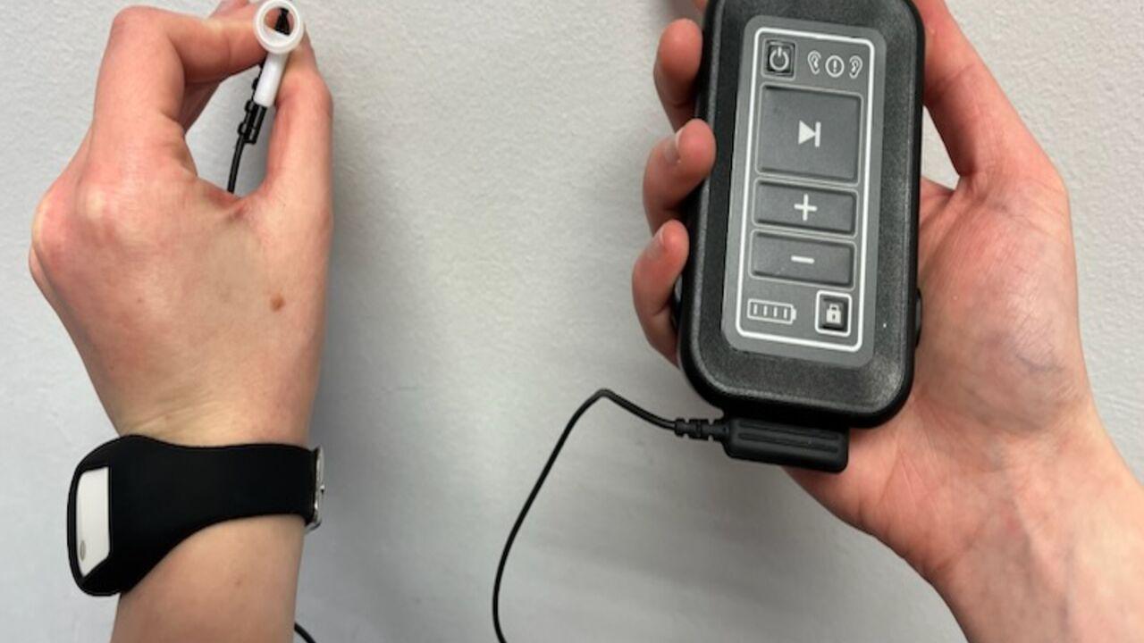 The device looks like a small, black remote with wires attached to an earpiece. The person holding the items is also wearing a black wristwatch.