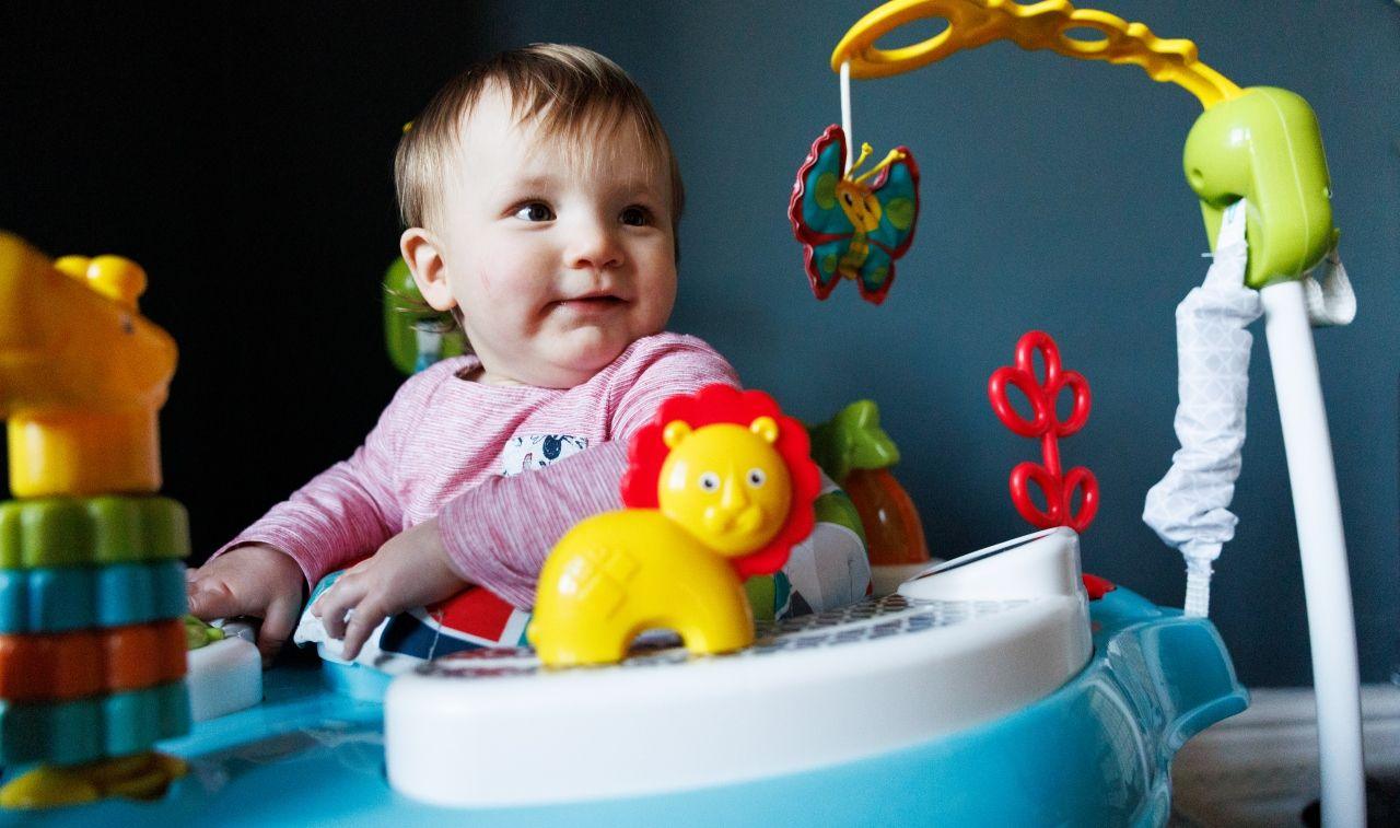 A baby girl wearing a pink top, surrounded by toys, sitting on a baby seat in a room with a blue wall.