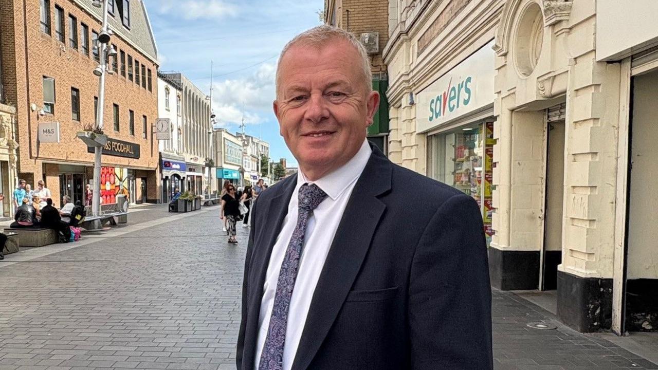 A man in his 60s, with short grey hair, smiles for the camera in a Grimsby shopping street. He is wearing a black suit.