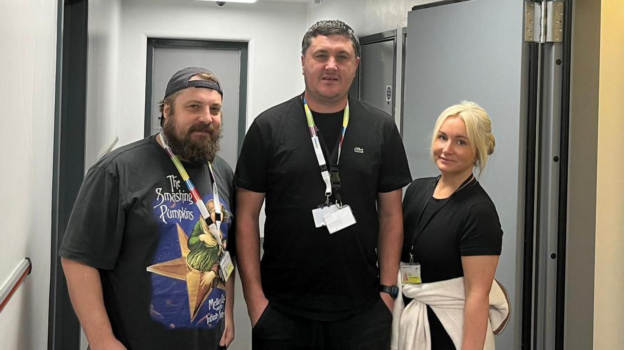 A man in a backwards cap and a Smashing Pumpkins t-shirt stands on the left of a tall man wearing two lanyards and a blonde-haired woman with a jacket tied round her waist on the right. They are standing in a custody suite with cell doors behind them and on the right.