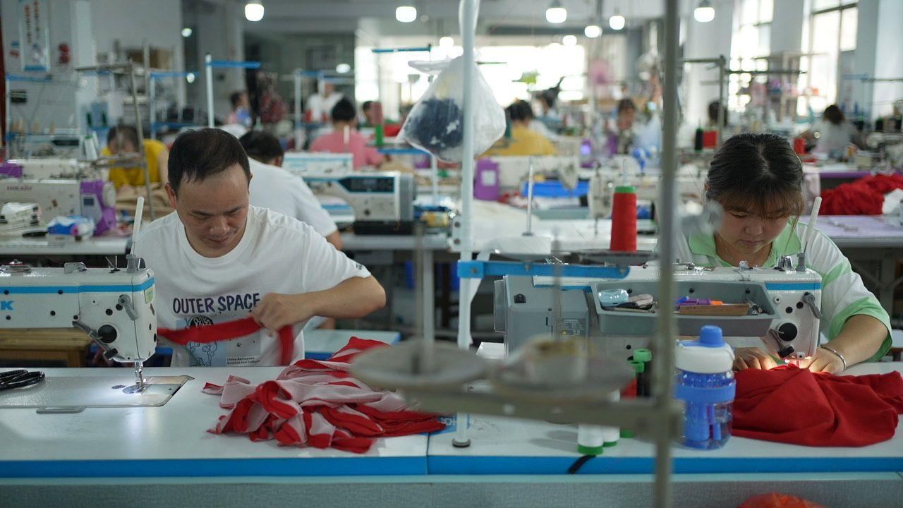 A man and a woman at a Shein factory, sitting side-by-side and working on sewing machines. They are cutting and sewing red fabric.   