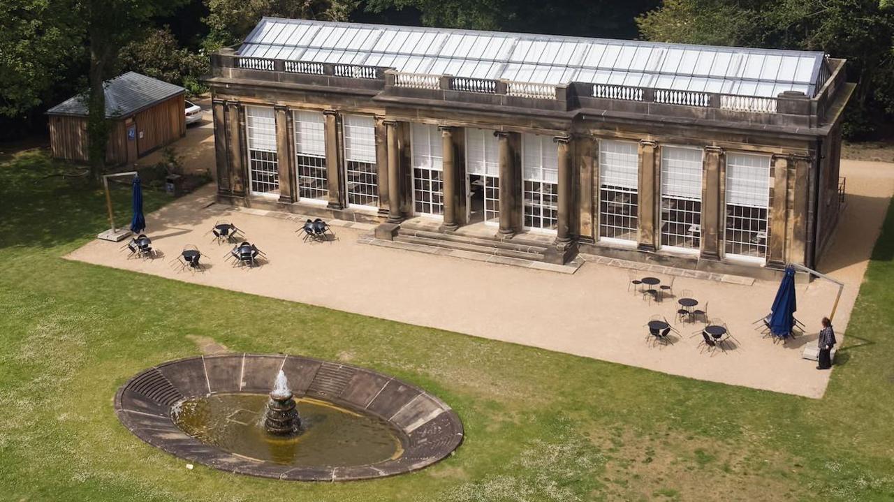 A single-storey historic building. Erected from sandstone with nine floor to ceiling large windows on the facade of the building. The Camellia House has a large fountain and lawns to the front, with woodland to the sides.