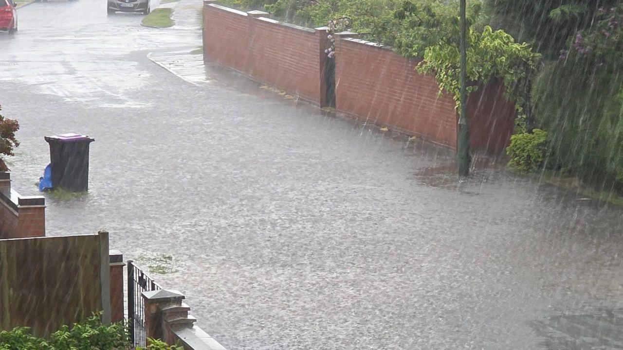 Rain hammers down on a road, submerging parts of it with water, between the fences of homes.