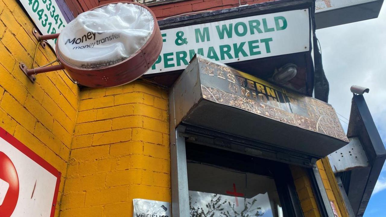 A corner shop with painted yellow brick walls. Signage around the entrance has been scorched and melted by fire. 