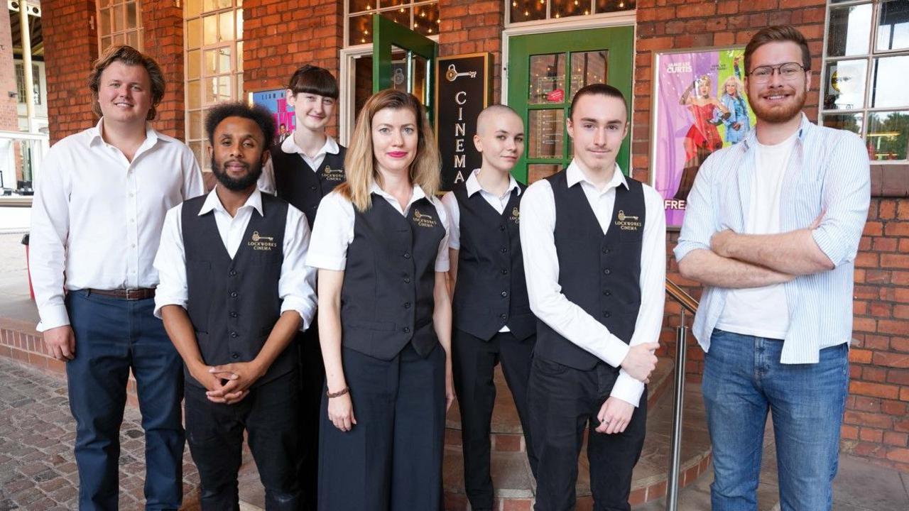 Seven people are standing outside a building with two green doors, one of which is open. Everyone, except the men on the far left and far right, are wearing black-and-white Lockworks Cinema uniforms that include its name.