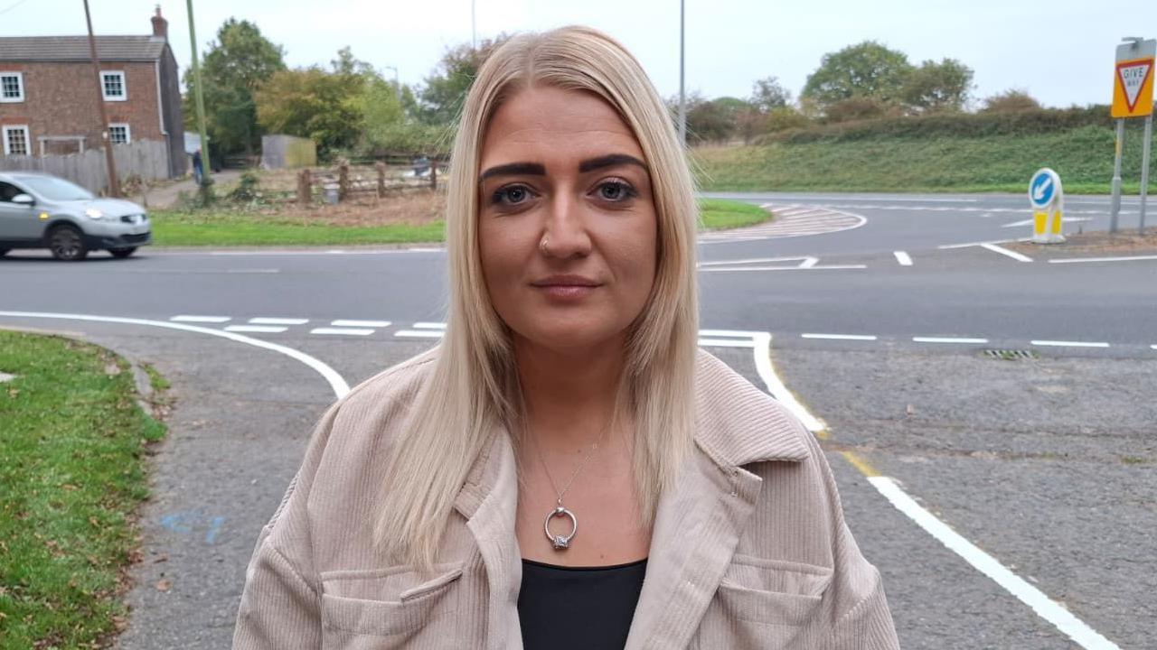 A woman with long blonde hair looks into the camera lens with a neutral expression on her face. She wears a cream-coloured jacket and black top, with a busy road junction in the background.