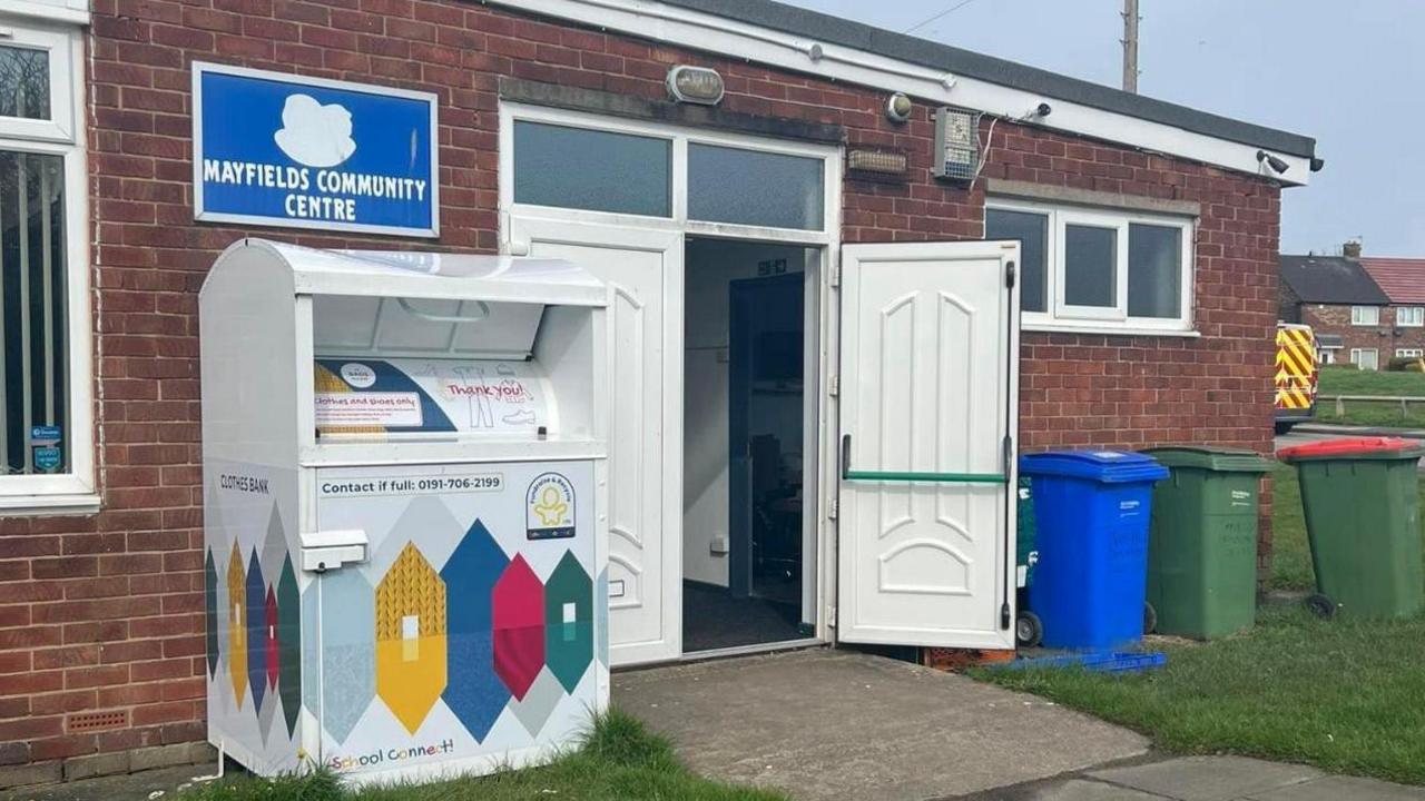 A single storey brick building with white double doors open and a sign on the wall saying Mayfields Community Centre. 