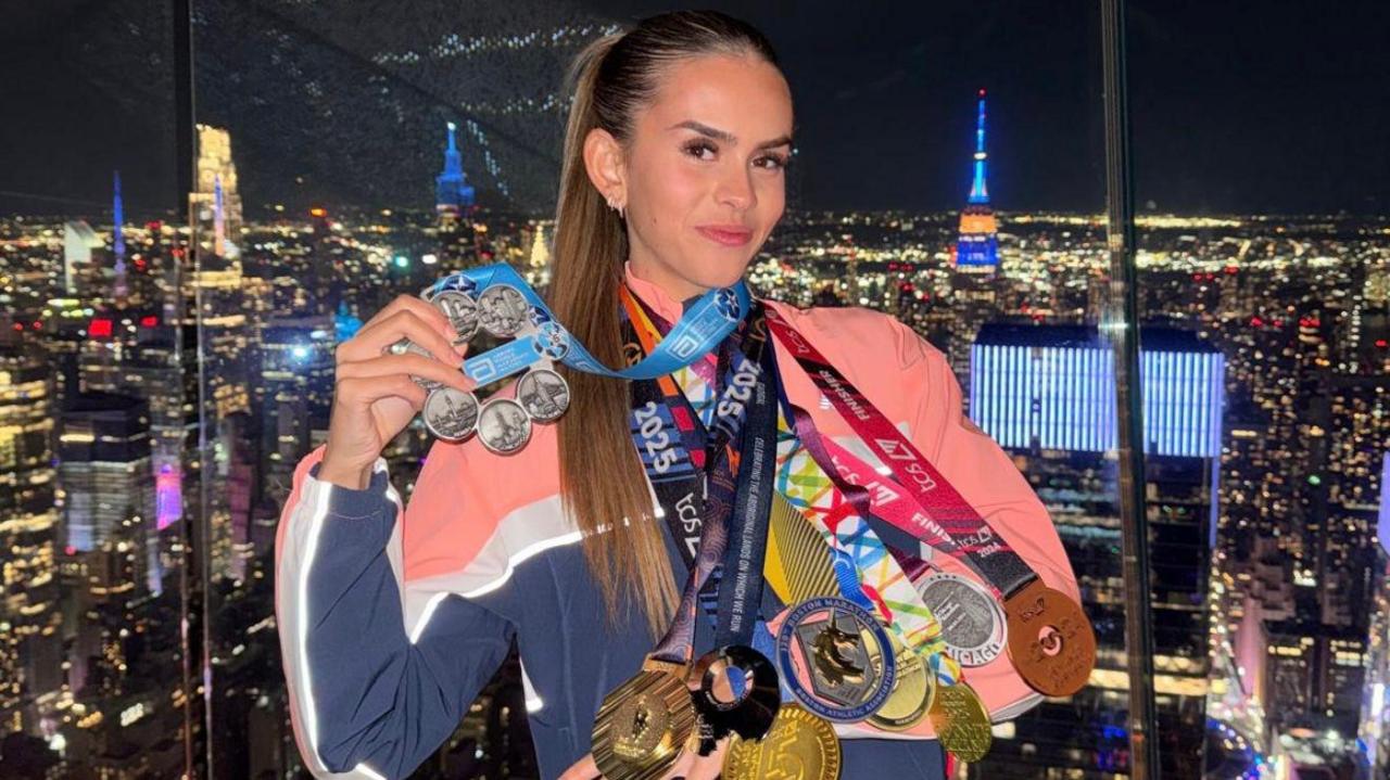 Lucy Scott has eight medals hanging from her neck. They vary from gold, silver and bronze with pink and blue coloured ribbons. She is stood in front of the view of New York City lit up at night. 