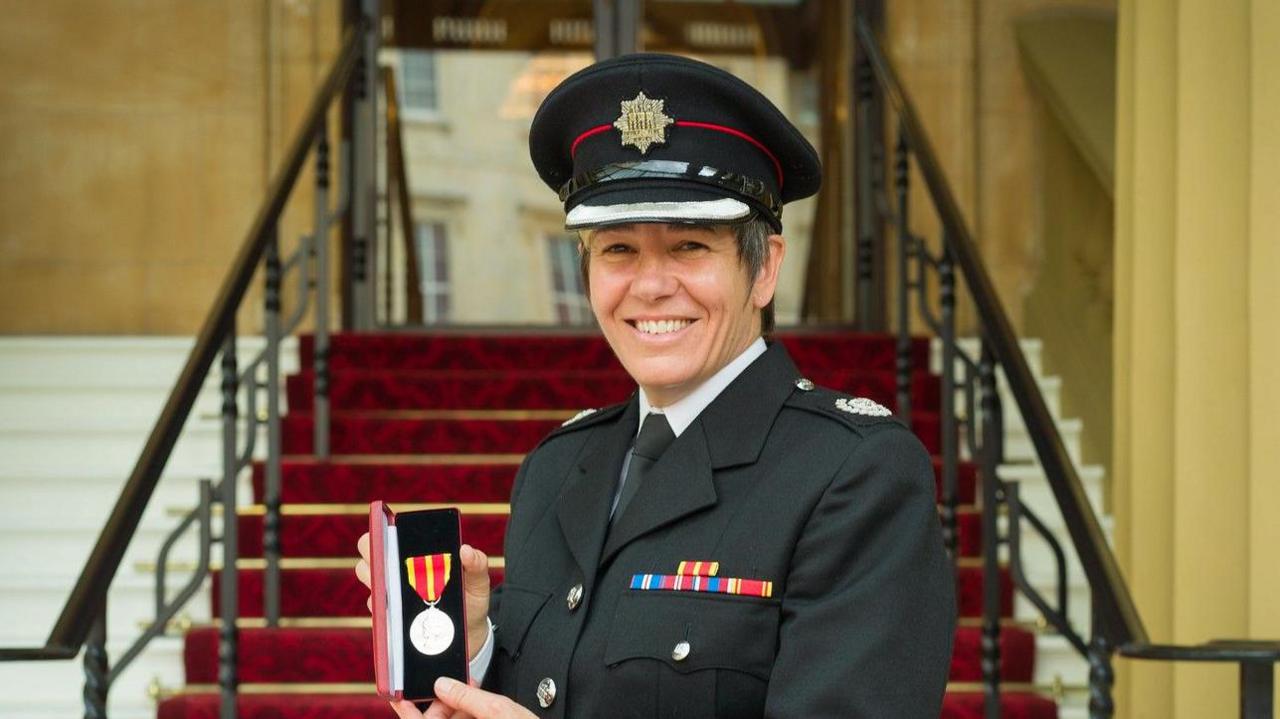 Ms Hammond smiles as she holds her Queen's Fire Service Medal. She wears a black uniform and a black hat. Stairs can be seen behind her.