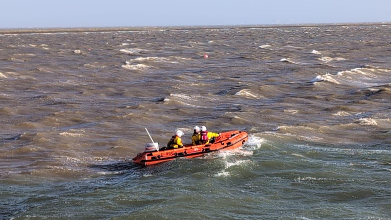 Three RNLI volunteers are sailing in an orange inflatable rescue boat in choppy waters. They are wearing yellow and red uniforms and white helmets. 