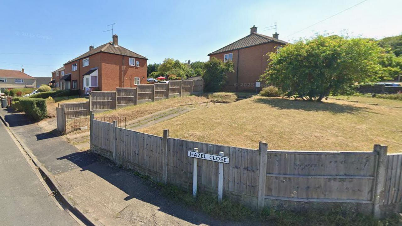 Hazel Close is a residential road with hedges and gardens and there are cars parked in driveways. The road sign is near a raised, fenced lawn, which has a tree in the middle of it.