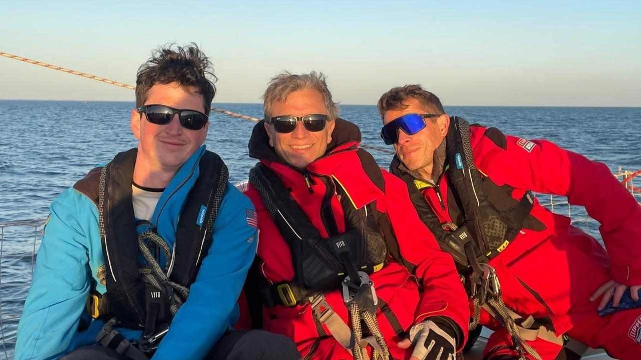 Kevin Lynch is far right of this three man photo. He is wearing a red full body sailing suit, with life-jacket and has sunglasses on. There is the ocean in the background.