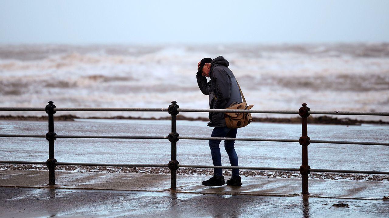 A person struggles against high winds while holding on to their hat in a winter coat, walking along a wet promenade with a stormy sea in the background, in New Brighton on 22 December.