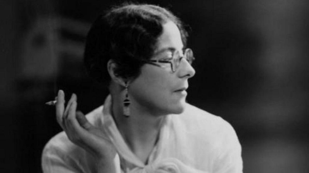 Black and white photo of Sylvia Townsend Warner smoking a cigarette. She is looking towards the right of the frame and holding the cigarette out to the left. She has dark hair, round glasses and pendant earrings.