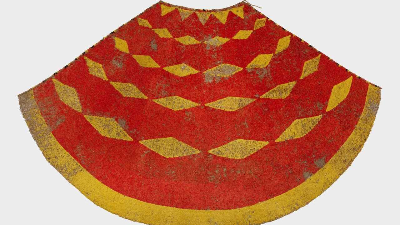 A 19th century yellow and red cloak made from feathers. It has an simple diamond shaped pattern and a yellow border. The feathers have fallen away in parts of the cloak.