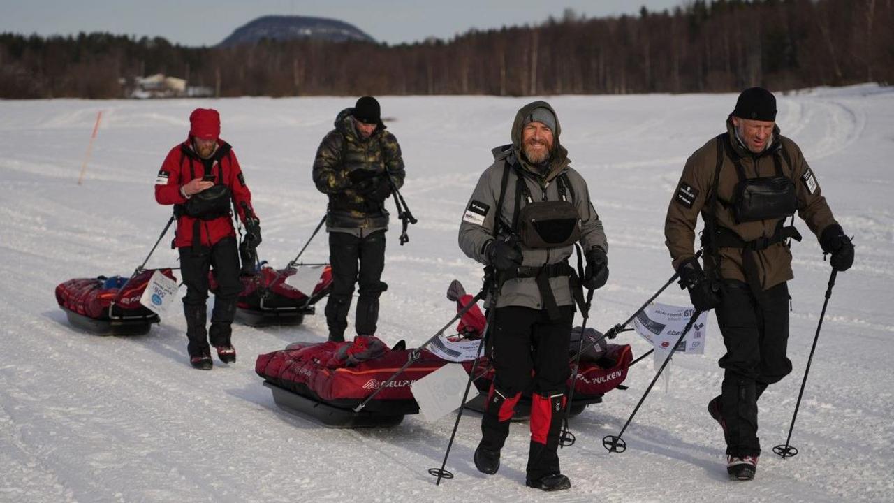 The group, pictured during the ultra-marathon, are walking in the snow near the camera. Trees are in the distance. They pull individual sledges and hold ski poles as they wear cold-weather clothing.