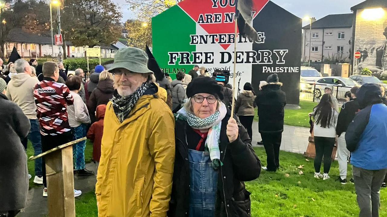 A number of people are standing in front of Free Derry Corner, a monument that was once a house's gable wall. The monument says 'You are now entering Free Derry'. It is usually white but at this time has been painted with the colours and design of the Palestinian flag. In front of the mural are dozens of people turned towards it. In the foreground of the picture are two people - a man and a woman. The man is on the left, he is wearing a green bucket hat and a yellow anorak. The woman is wearing a black coat, scarf and black hat. She is holding up a black flag on a flagpole, to commemorate those who died on Bloody Sunday.