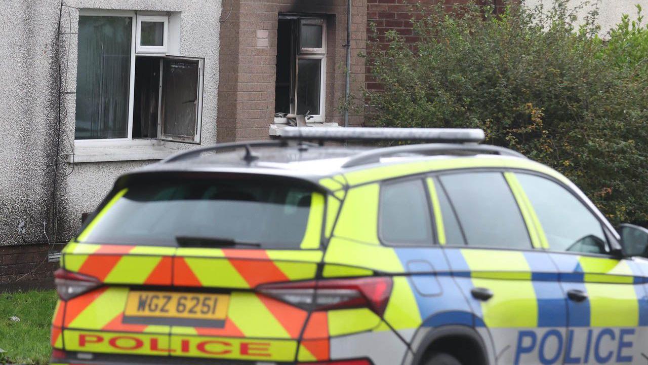 A police car with yellow and orange stripes sits in front of a brown brick building with black burn marks around the windows.