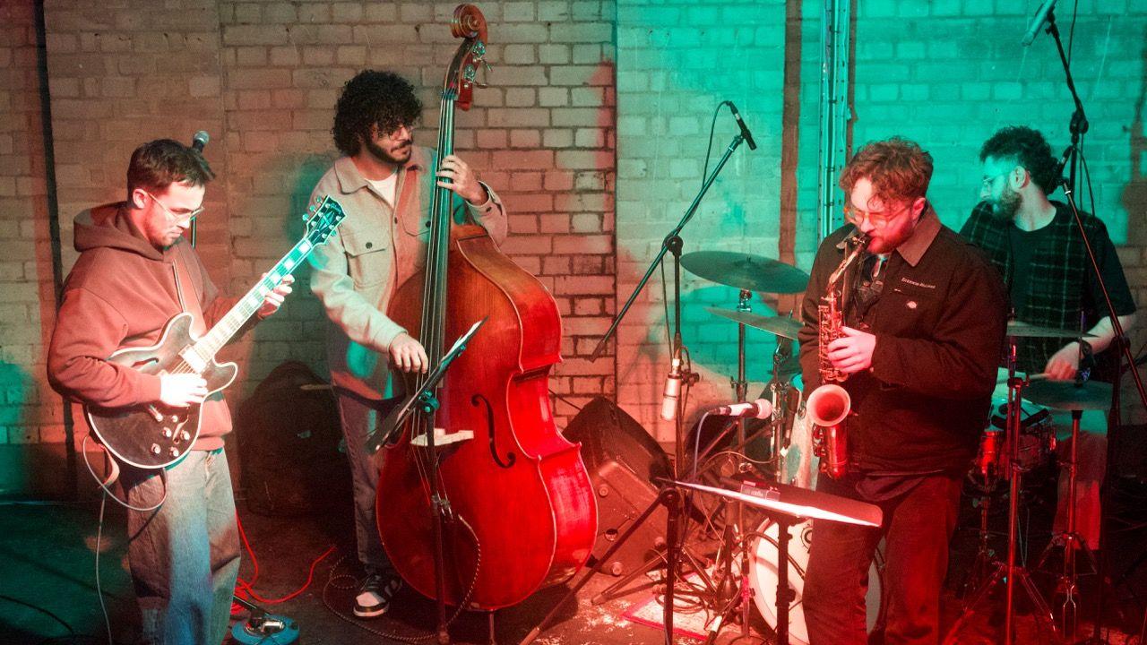 Four men are wearing dark coloured clothing and standing on a stage. They are playing instruments including the double bass, guitar, saxophone and drums. Green and red neon lights are shining on them